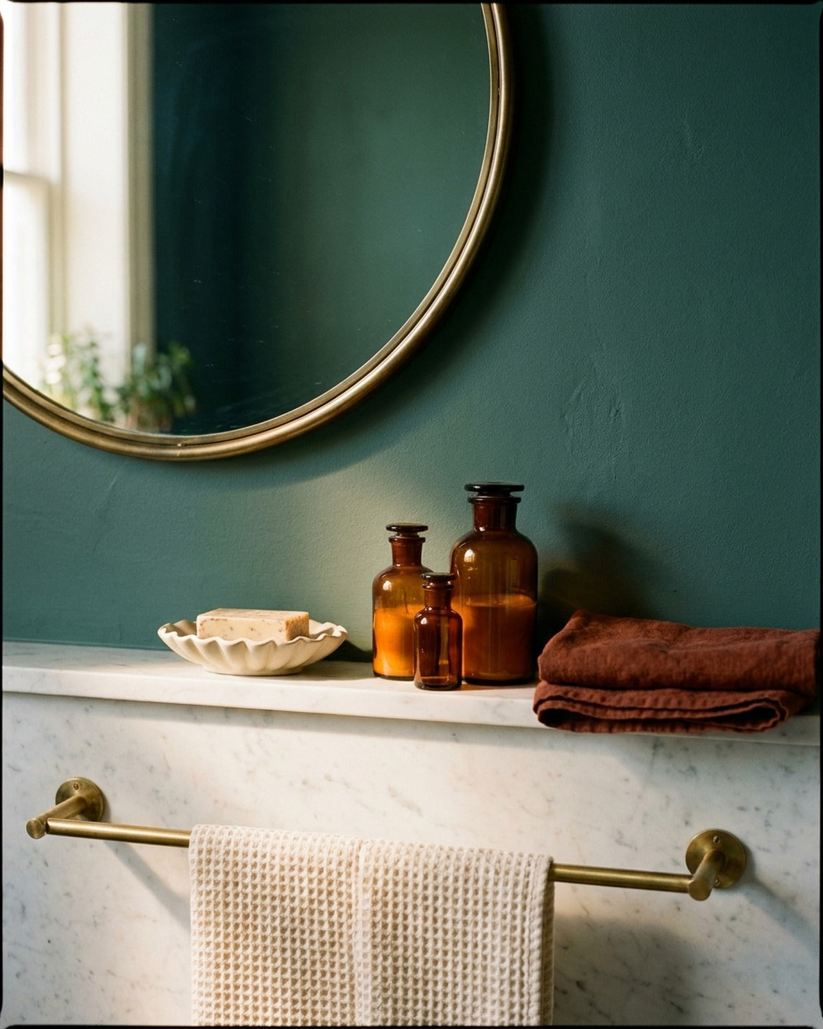 A round brass mirror above a marble ledge with a scalloped soap dish, amber glass bottles and a rust linen towel on a deep teal wall