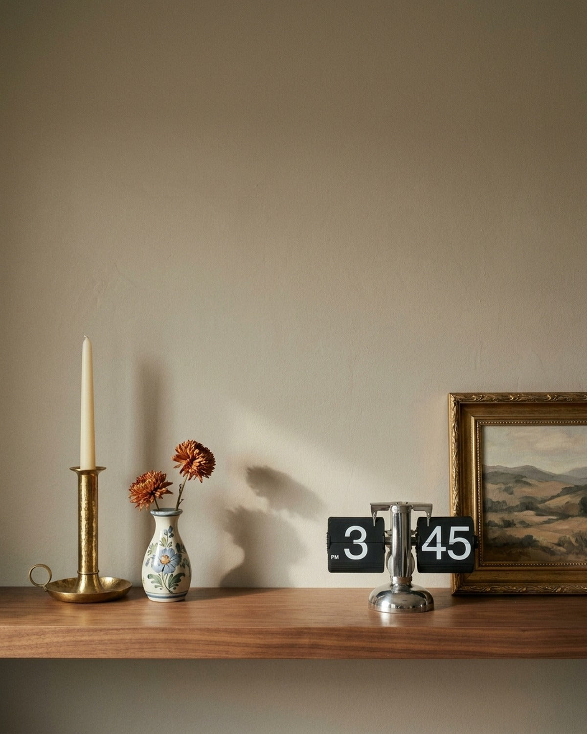 A brass candlestick, floral bud vase and retro flip clock on a walnut shelf against a warm taupe wall