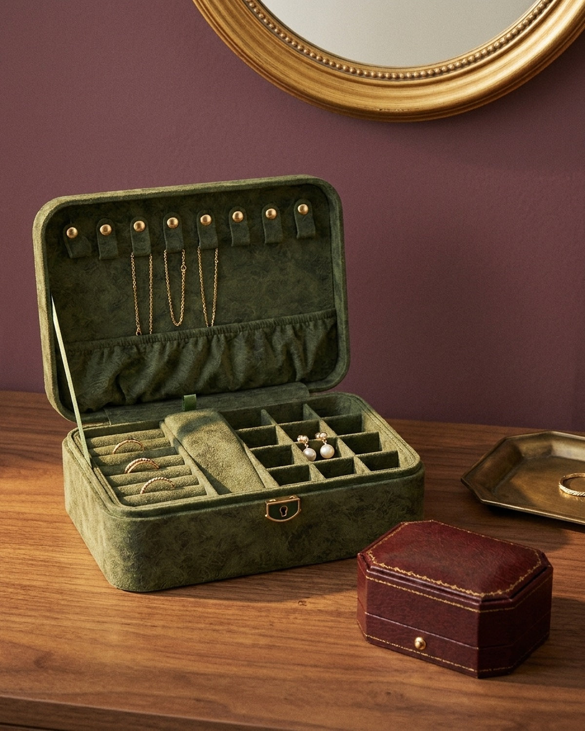 An open olive suede jewellery case with gold rings and pearl earrings on a walnut dressing table against a plum wall
