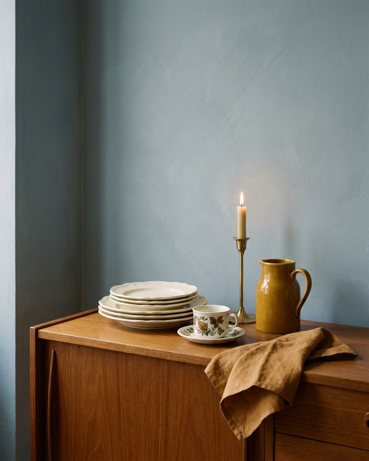 Cream scalloped plates, a mustard jug and a brass candlestick on a walnut dresser against a chalky blue-grey wall