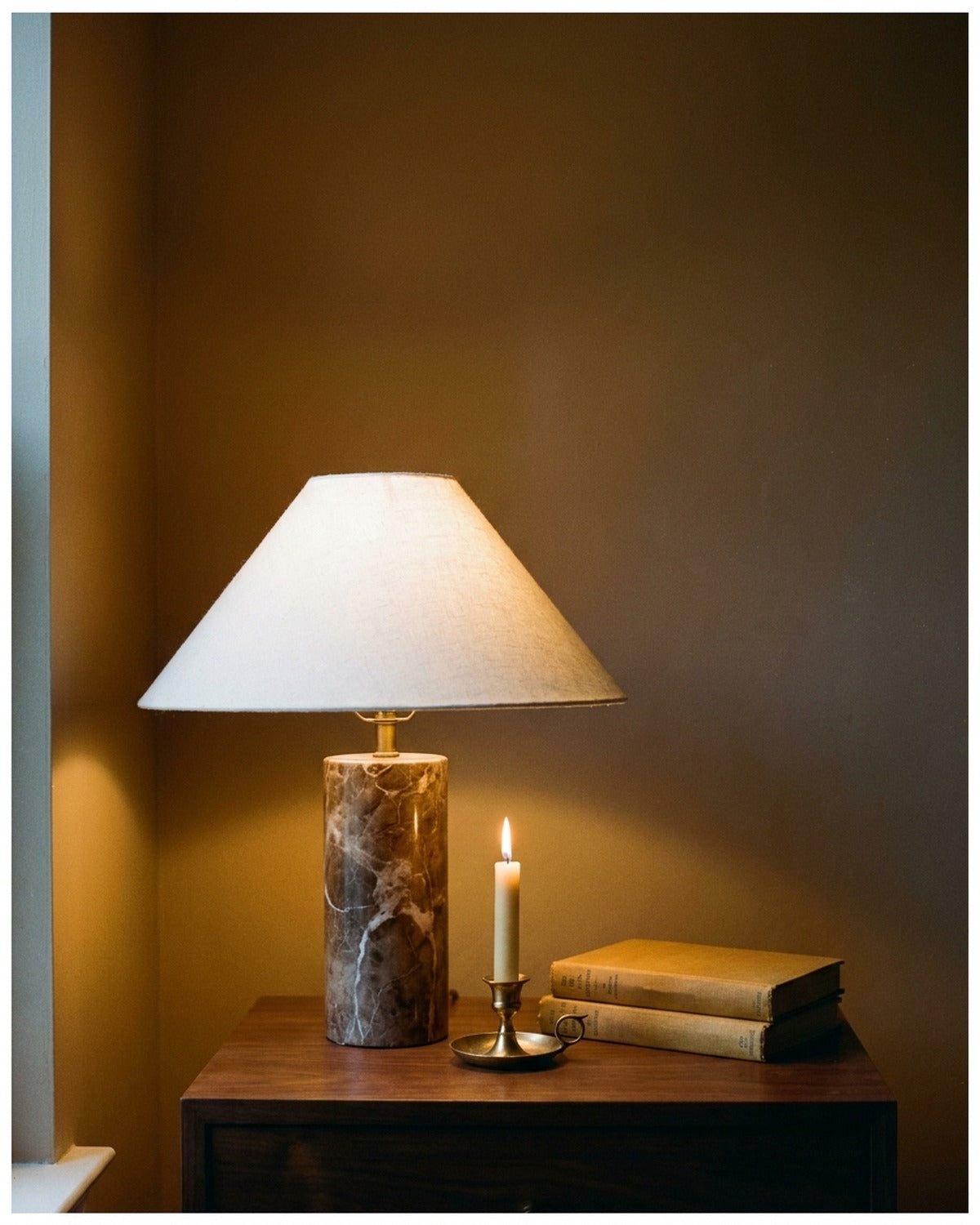 A lit marble table lamp and brass candlestick on a walnut side table against a deep ochre wall