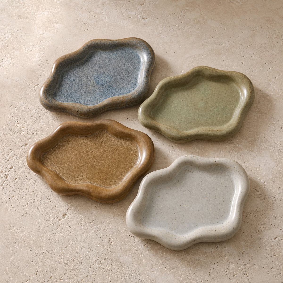 Four cloud-shaped ceramic catchall trays in bronze brown, olive green, blue gray and speckled ivory on warm limestone.