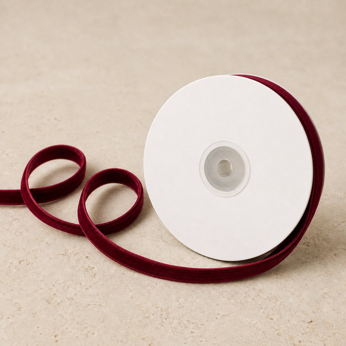 Wine Burgundy double-sided velvet ribbon unwinding continuously from a plain white spool on warm limestone.