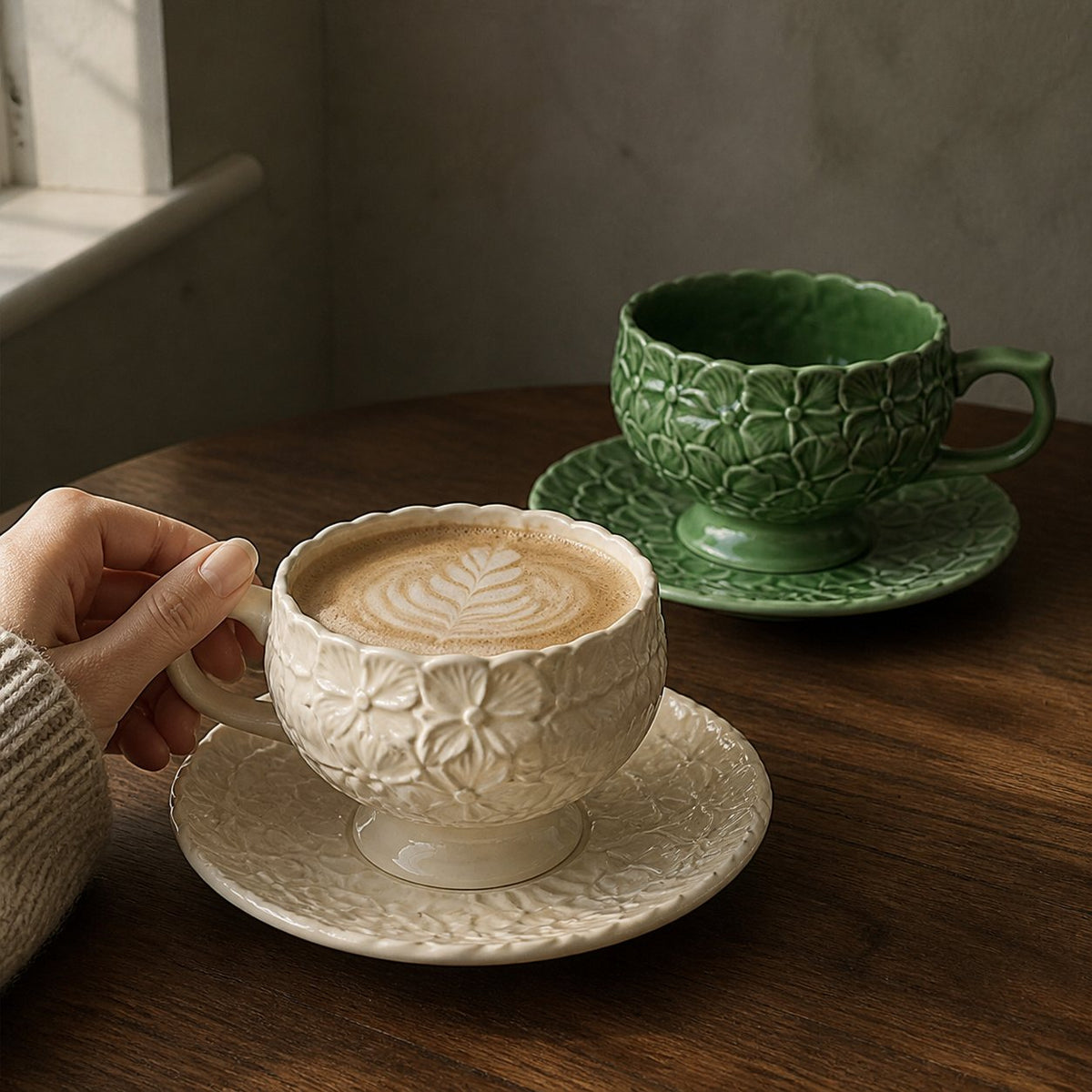Hand holding an Ivory Bloom hydrangea relief cup filled with coffee beside the Sage Garden set.