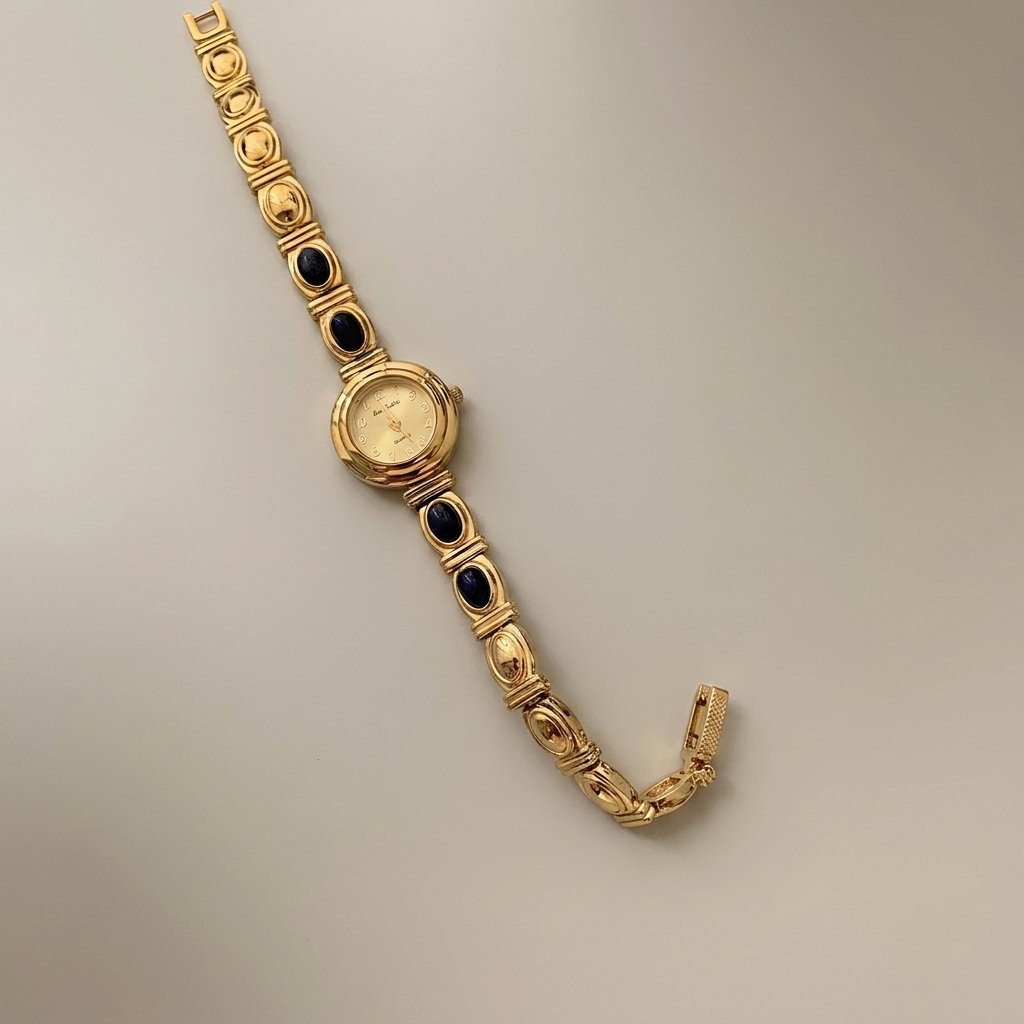 A vintage gold-tone watch with a rectangular link bracelet featuring dark oval inlays, displayed diagonally on a neutral light gray background.