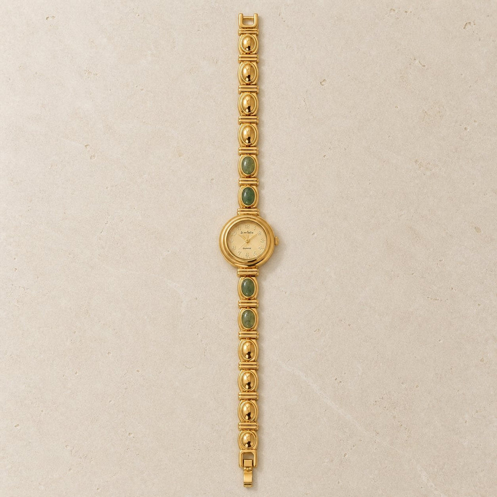 Full-length gold-tone alloy bracelet watch with four green oval inlays laid vertically on a pale stone background.