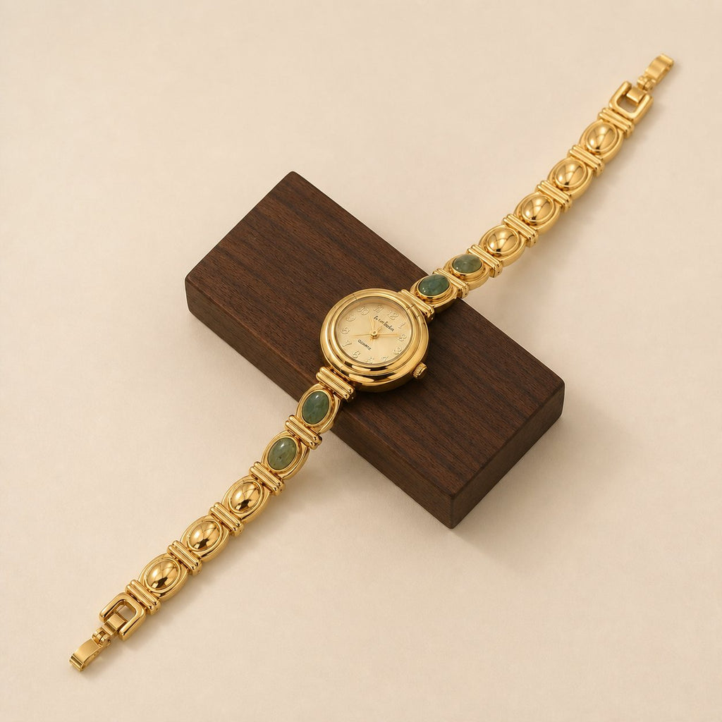 Gold-tone alloy bracelet watch with four green oval inlays displayed diagonally across a dark walnut block.