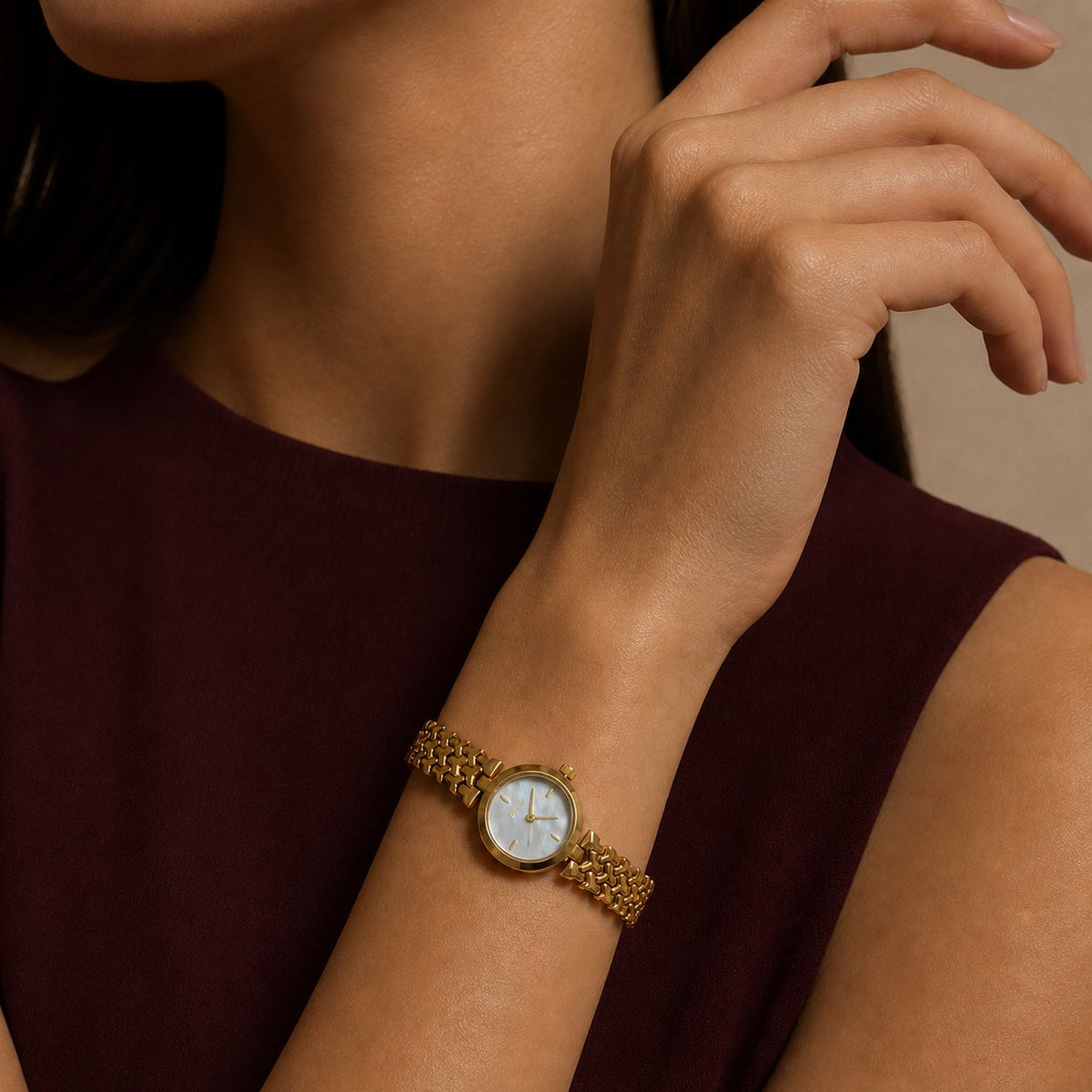 Woman wearing a gold-tone woven-link bracelet watch with a burgundy sleeveless top in an editorial studio portrait.