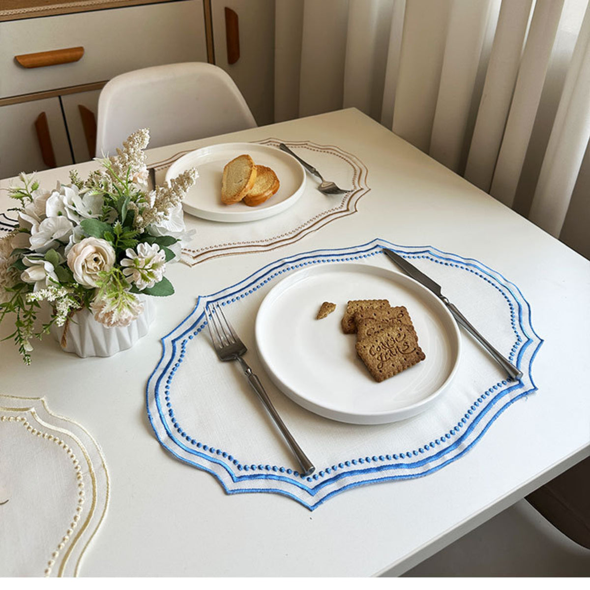 Camel-trim and sky-blue-trim placemats set with breakfast plates on a bright dining table with fresh flowers