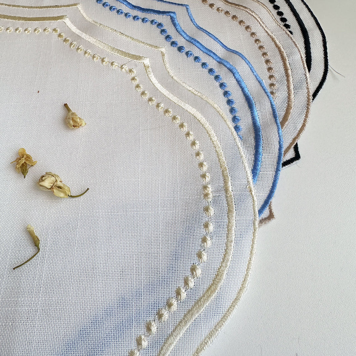 Close-up of the ivory linen-look fabric weave showing all four embroidered trim colors side by side — black, sky blue, camel, and cream