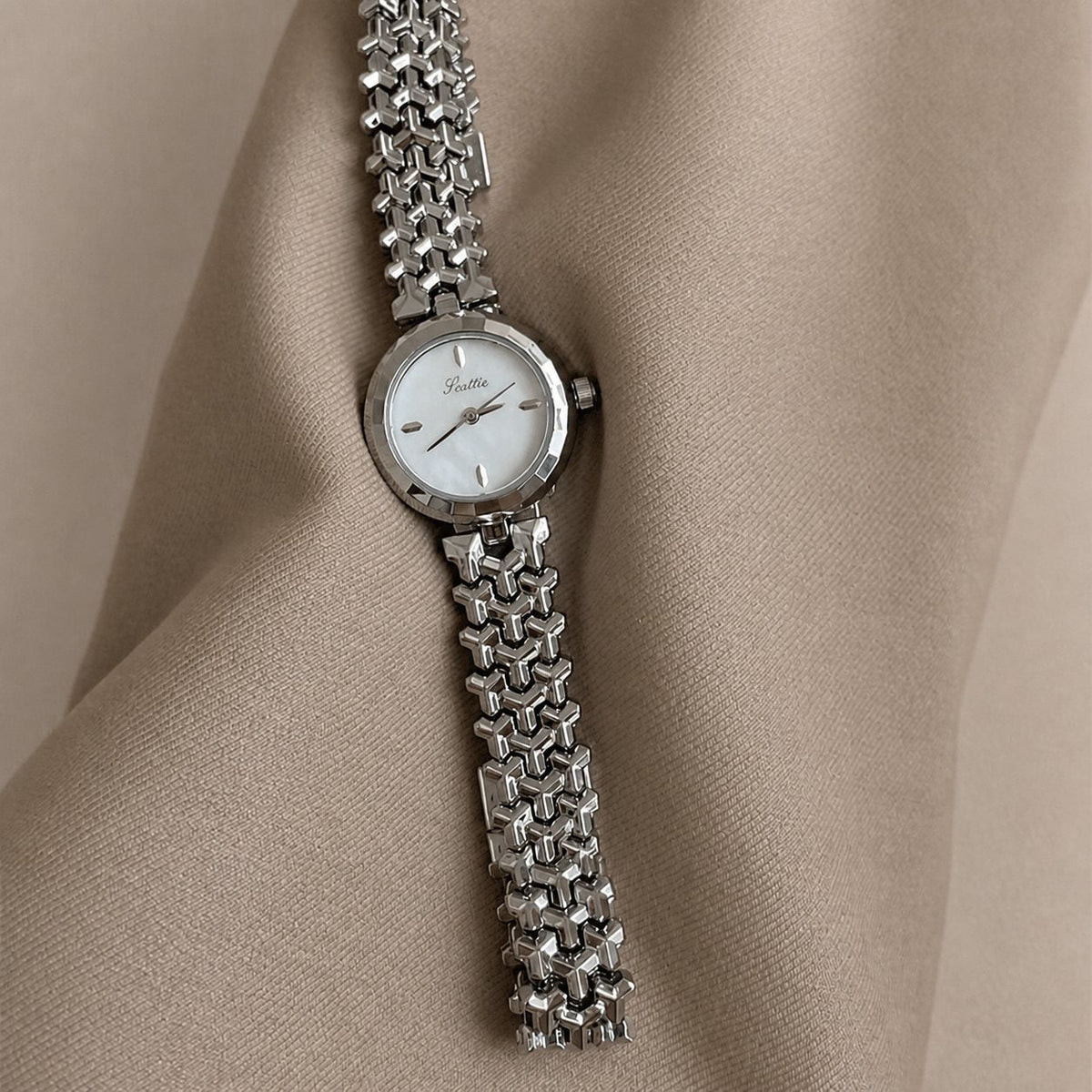 Silver-tone woven-link bracelet watch arranged vertically on warm beige fabric folds.